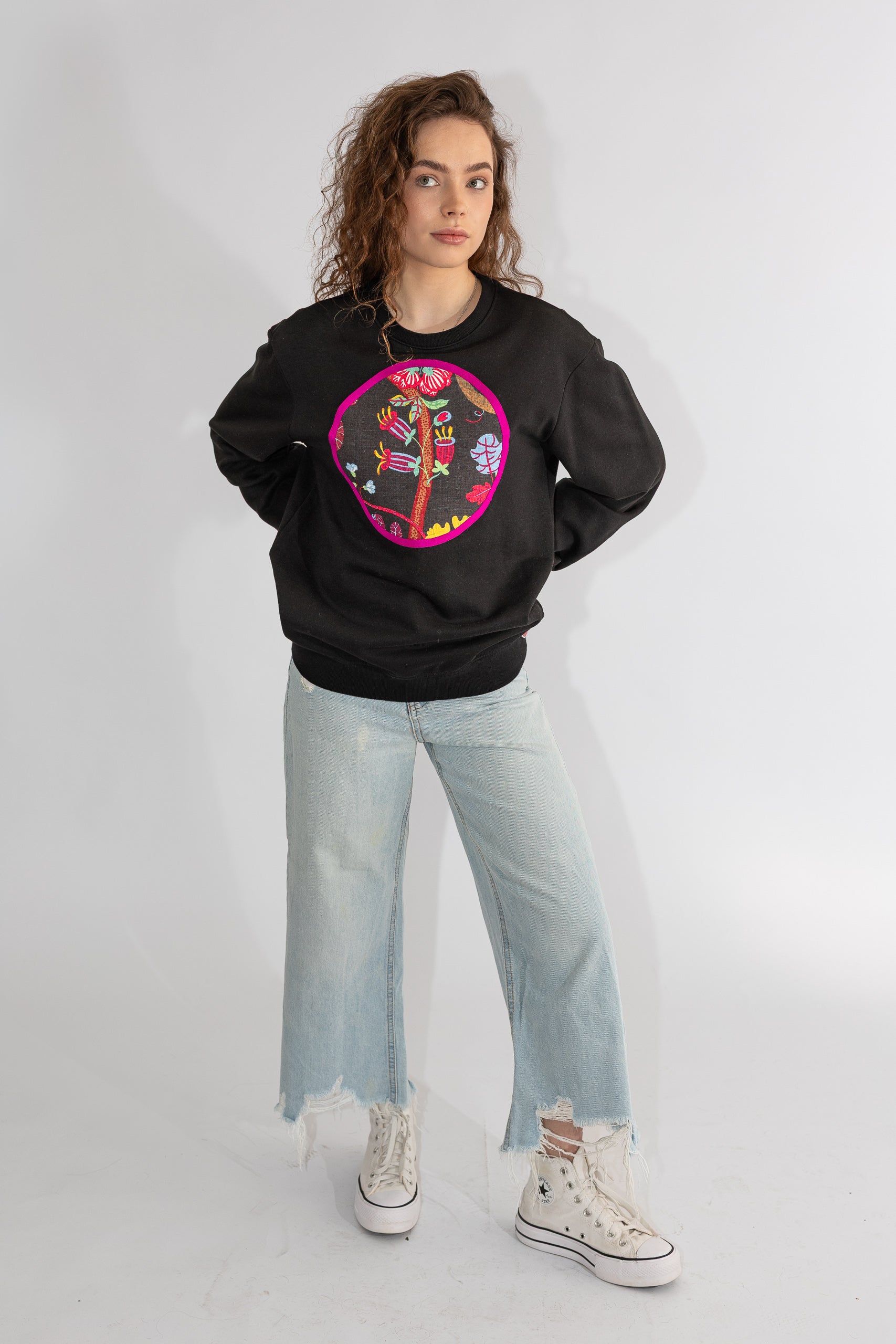 Person wearing a black hoodie with a colorful design, light blue jeans, and white sneakers on a white background