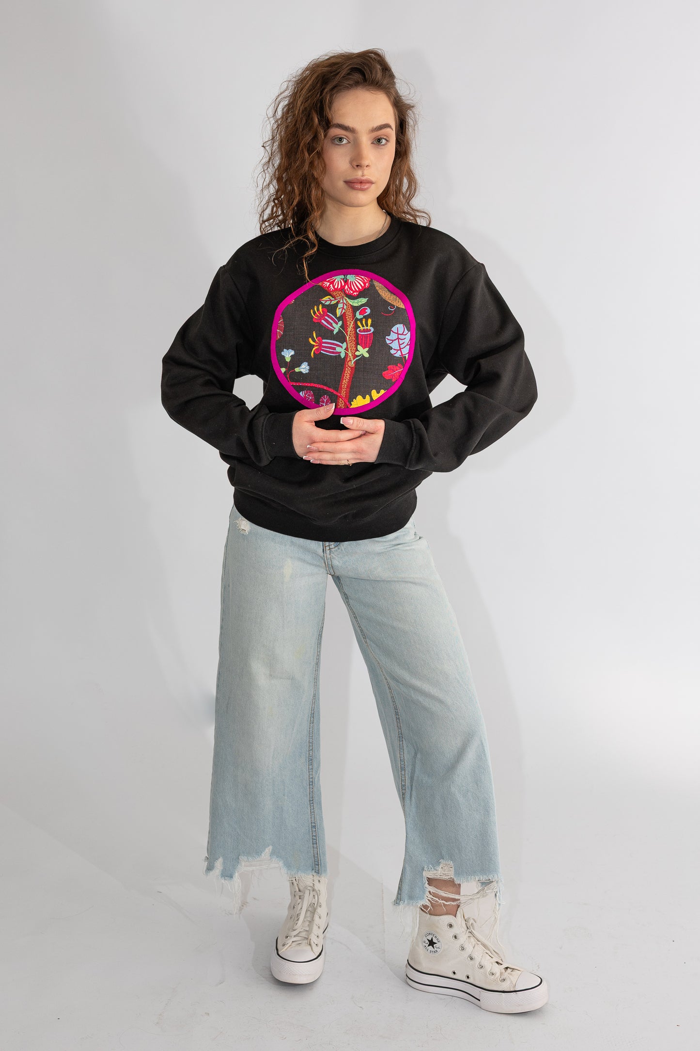 Person wearing a black sweatshirt with a colorful design, light blue jeans, and white sneakers on a plain background